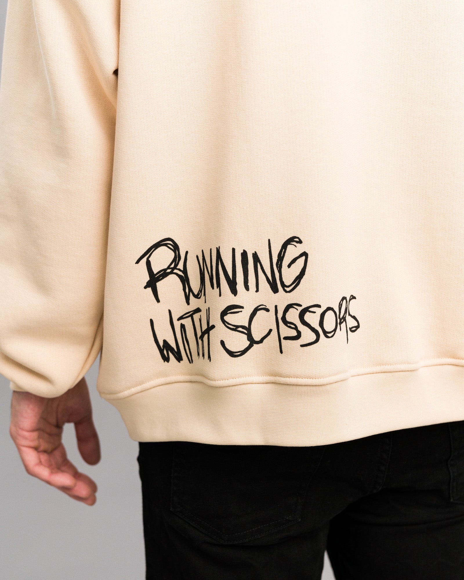 Detailansicht des Rückendrucks am Hoodie: Schwarzer Graffiti-Schriftzug „RUNNING WITH SCISSORS“ auf beigefarbenem Stoff, unten rechts platziert.