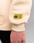 Nahaufnahme des gelben Labelpatches am rechten Ärmelbündchen des Hoodies mit schwarzem Text und Symbol, der technische Codes und ein Logo zeigt.