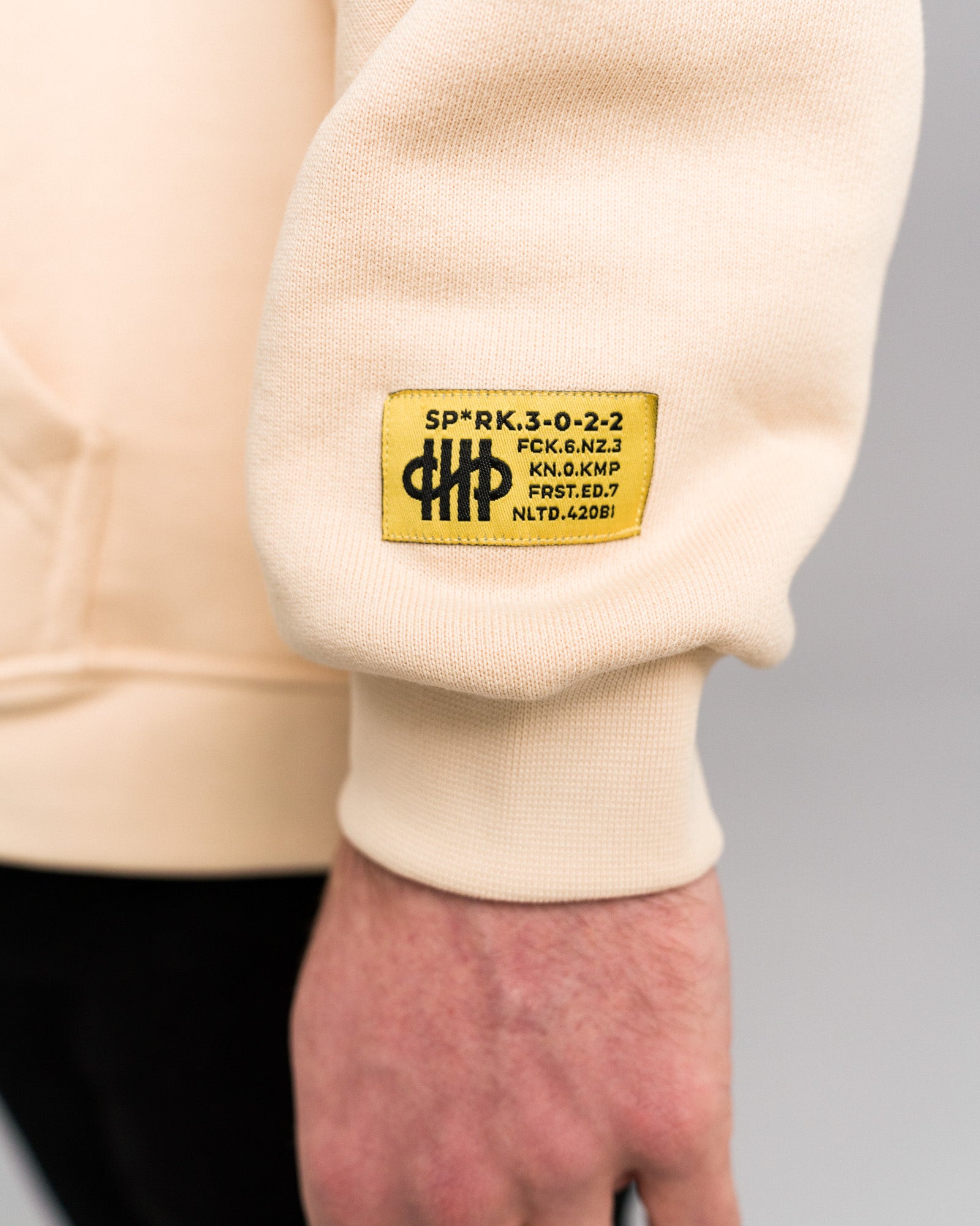 Nahaufnahme des gelben Labelpatches am rechten Ärmelbündchen des Hoodies mit schwarzem Text und Symbol, der technische Codes und ein Logo zeigt.