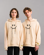 Bild 6:
Zwei Personen tragen identische beigefarbene Hoodies mit dem tropfenden Smiley-Print auf der Vorderseite. Beide Modelle zeigen die Labelapplikation am rechten Ärmel.


