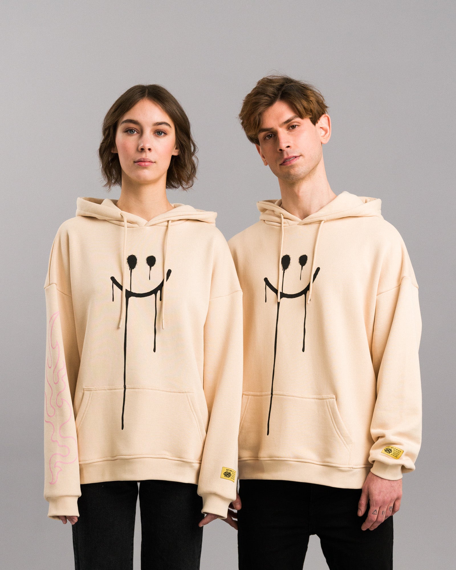 Bild 6:
Zwei Personen tragen identische beigefarbene Hoodies mit dem tropfenden Smiley-Print auf der Vorderseite. Beide Modelle zeigen die Labelapplikation am rechten Ärmel.

