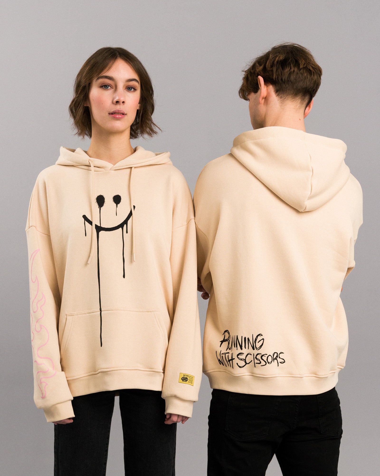 Zwei Personen in frontaler und rückwärtiger Ansicht des Hoodies. Die Vorderseite zeigt den schwarzen Smiley, die Rückseite den Schriftzug „RUNNING WITH SCISSORS“.