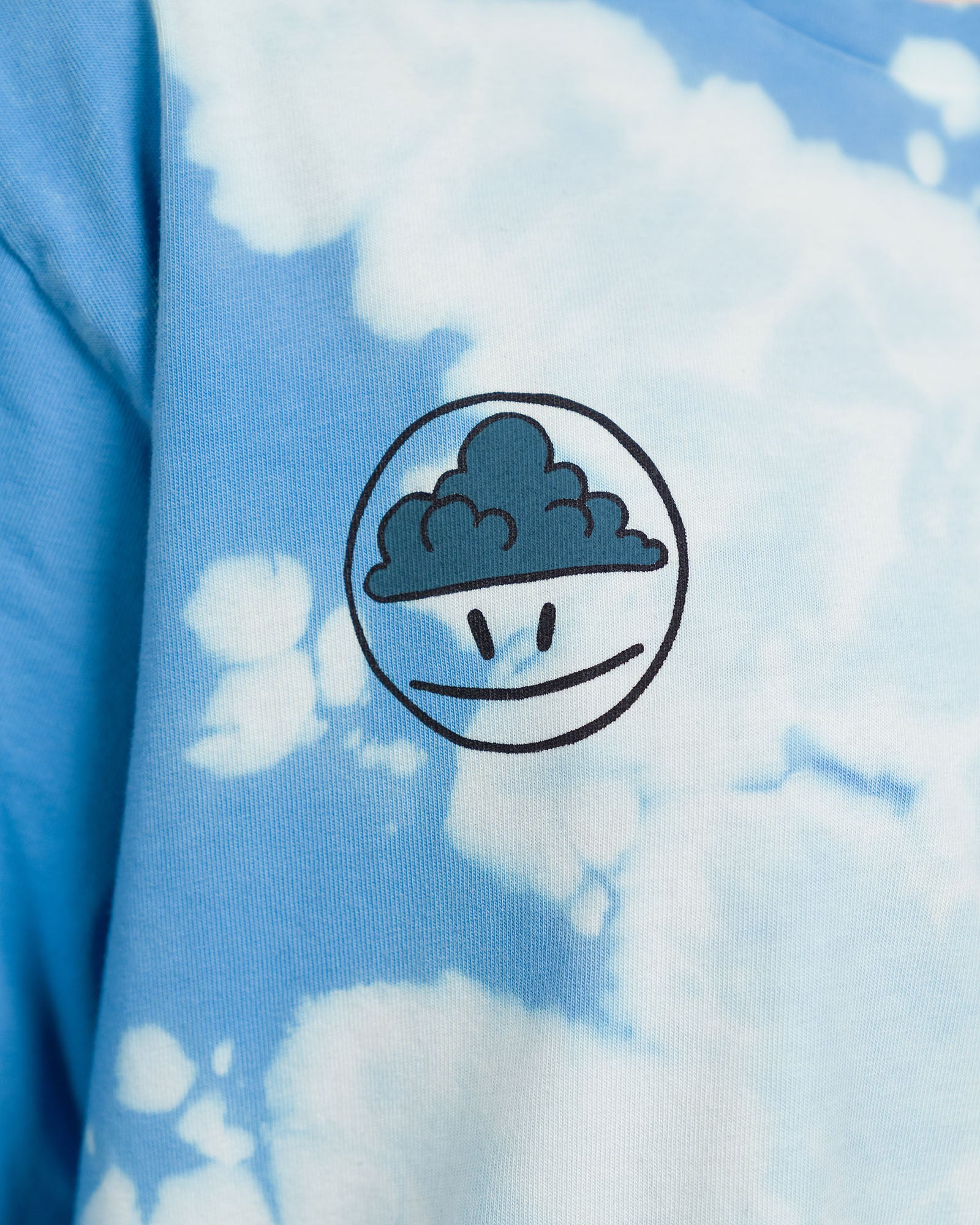 Detailansicht des Smiley-Prints auf dem T-Shirt mit Batikmuster. Das Motiv zeigt ein rundes Gesicht mit ernster Miene und einer dunklen Wolke im oberen Bereich des Kopfes.