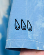Detailaufnahme des rechten Ärmels eines Batikshirts. Drei aufgestickte schwarze Tropfenmotive mit lächelnden Gesichtern sind nebeneinander in vertikaler Anordnung zu sehen.