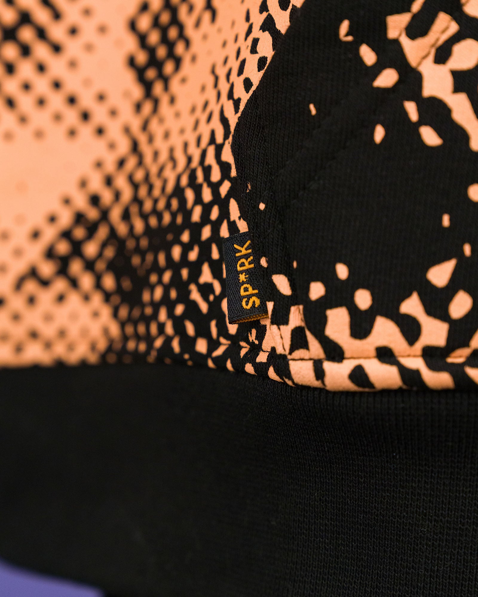 Detailaufnahme eines kleinen, schwarzen Weblabels mit gelber Aufschrift „SP*RK“, das seitlich in die Naht auf Hüfthöhe eingenäht ist. Der umgebende Stoff zeigt ein Muster aus orangefarbenen, organischen Rasterpunkten auf schwarzem Grund.

