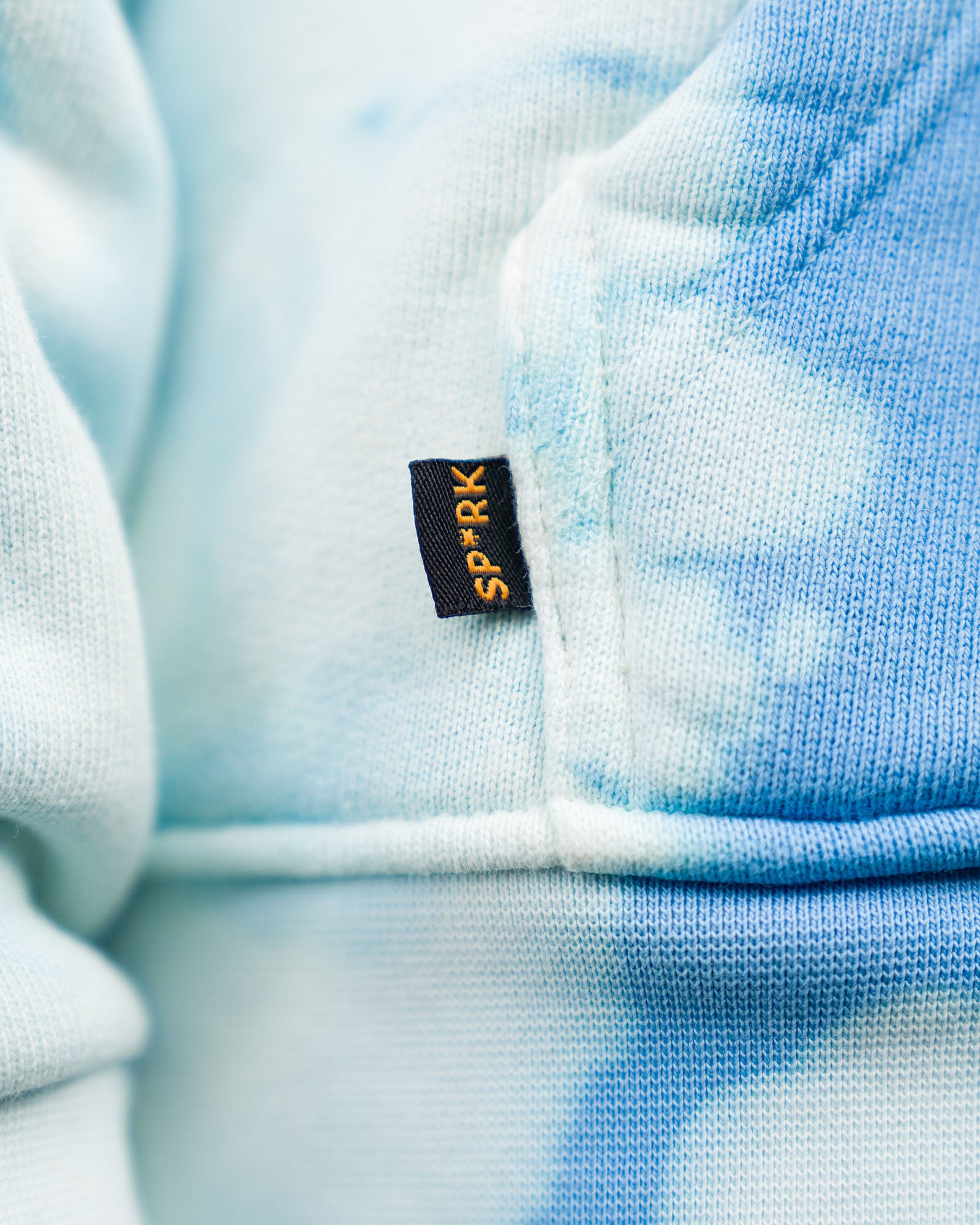 Detailaufnahme eines kleinen Stofflabels an der Seitennaht der Kängurutasche mit orangem SP*RK-Schriftzug auf dunkelblauem Etikett.